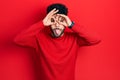 Young arab man with beard wearing casual red sweater doing ok gesture like binoculars sticking tongue out, eyes looking through Royalty Free Stock Photo