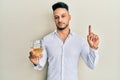 Young arab man drinking a glass of white wine smiling with an idea or question pointing finger with happy face, number one Royalty Free Stock Photo