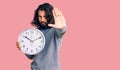 Young arab man holding big clock with open hand doing stop sign with serious and confident expression, defense gesture Royalty Free Stock Photo