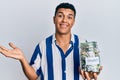 Young arab man holding charity jar with money celebrating achievement with happy smile and winner expression with raised hand Royalty Free Stock Photo