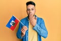 Young arab man holding haiti flag covering mouth with hand, shocked and afraid for mistake