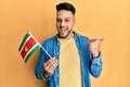 Young arab man holding suriname flag pointing thumb up to the side smiling happy with open mouth