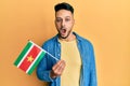 Young arab man holding suriname flag scared and amazed with open mouth for surprise, disbelief face