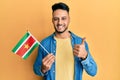 Young arab man holding suriname flag smiling happy and positive, thumb up doing excellent and approval sign