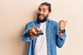 Young arab man holding television remote control pointing thumb up to the side smiling happy with open mouth Royalty Free Stock Photo