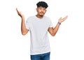 Young arab man wearing casual white t shirt clueless and confused expression with arms and hands raised