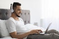 Young Arab Man Wearing Wireless Headphones Using Laptop While Relaxing In Bed Royalty Free Stock Photo