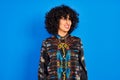 Young arab woman with curly hair wearing colorful shirt over isolated blue background looking away to side with smile on face, Royalty Free Stock Photo