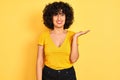 Young arab woman with curly hair wearing t-shirt standing over isolated yellow background smiling cheerful presenting and pointing Royalty Free Stock Photo
