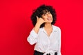 Young arab woman with curly hair wearing white casual shirt over isolated red background smiling doing phone gesture with hand and Royalty Free Stock Photo