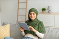 Young Arab woman in hijab using digital tablet for work, communication or studies on cozy couch at home