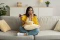 Young arab woman sitting on couch with TV remote, choosing movie to watch and eating popcorn, enjoying free time Royalty Free Stock Photo