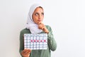Young Arab woman wearing hijab showing menstruation calendar over isolated background serious face thinking about question, very