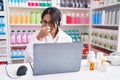 Young arab woman working at pharmacy drugstore using laptop smelling something stinky and disgusting, intolerable smell, holding