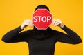 Young arabian muslim woman in hijab black clothes hold in hands red sign with Stop title isolated on yellow wall