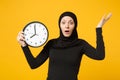 Young arabian muslim woman in hijab black clothes hold in hands round clock isolated on yellow wall background studio