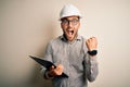 Young architect inspector man wearing builder safety helmet and using clipboard screaming proud and celebrating victory and Royalty Free Stock Photo