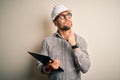 Young architect inspector man wearing builder safety helmet and using clipboard serious face thinking about question, very