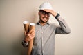 Young architect man wearing contractor helmet holding project paper plan over isolated background stressed with hand on head, Royalty Free Stock Photo