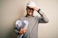 Young architect man wearing contractor helmet holding project paper plan over isolated background stressed with hand on head, Royalty Free Stock Photo