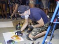 A young artist paints a picture of multicolored spray paint at night in front of everybody. A young guy in a respirator.