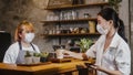 Young Asia female barista wear face mask serving take away hot coffee paper cup to consumer at cafe. Owner small business, Royalty Free Stock Photo