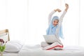 Young asian arab woman using laptop sitting on bed in bedroom. Beautiful muslim girl in sleepwear with blue hijab stretching her Royalty Free Stock Photo