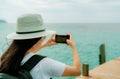 Young Asian backpacker woman wear hat use smartphone taking photo at pier. Summer vacation at tropical paradise beach. Happy Royalty Free Stock Photo