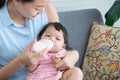 Young Asian beautiful mother smiling, holding and feeding milk from bottle to cute little Caucasian 7 months newborn baby, Royalty Free Stock Photo