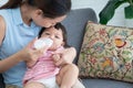 Young Asian beautiful mother smiling, holding and feeding milk from bottle to cute little Caucasian 7 months newborn baby,