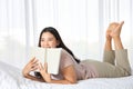 Young Asian brunette woman lying down on the bed while reading a book in cozy and relax feeling Royalty Free Stock Photo