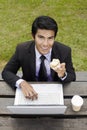 Young Asian businessman having a light snack in a park with his laptop. Conceptual image Royalty Free Stock Photo