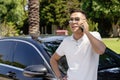 A young Asian businessman, a man in a white shirt and sunglasses, stands near a modern car and talks on a mobile phone, solves