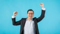 Young Asian businessman wearing glasses rise hand up and feeling happy about his business success idea isolated on blue background Royalty Free Stock Photo