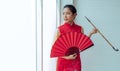 Young Asian Chinese beautiful woman wearing red traditional dress, holding folding fan and pipe, standing beside window in a day