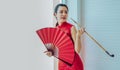 Young Asian Chinese beautiful woman wearing red traditional dress, holding folding fan and pipe, standing beside window in a day