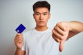 Young asian chinese man holding credit card standing over isolated white background with angry face, negative sign showing dislike Royalty Free Stock Photo