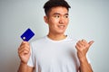 Young asian chinese man holding credit card standing over isolated white background pointing and showing with thumb up to the side Royalty Free Stock Photo