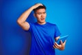 Young asian chinese man using tablet standing over isolated blue background stressed with hand on head, shocked with shame and Royalty Free Stock Photo