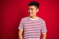 Young asian chinese man wearing striped t-shirt standing over isolated red background looking away to side with smile on face, Royalty Free Stock Photo