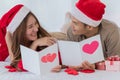 Young asian couple wearing santa hat lay down on white sheet bed together while woman smiling and writing card with white pen and Royalty Free Stock Photo