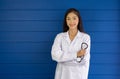 Young asian doctor in white gown and stethoscope stand with arm cross in front of blue wall