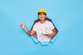 Young asian engineer in yellow helmet holds white roll paper projects smiles and gesturing thumbs up in blue background Royalty Free Stock Photo