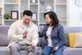 Young Asian family sitting on sofa at home. A man holds his hand to his chest, heart attack, stroke, panic attack, feels Royalty Free Stock Photo