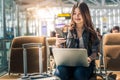 Young Asian female passenger using laptop and smart phone while sitting on seat in terminal hall and waiting for flight in airport Royalty Free Stock Photo