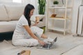 Young Asian female student looking at laptop screen sitting on floor at home with notebooks to write, lifestyle work