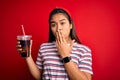 Young asian girl drinking cola fizzy refreshment using straw over isolated red background cover mouth with hand shocked with shame Royalty Free Stock Photo