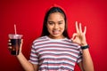 Young asian girl drinking cola fizzy refreshment using straw over isolated red background doing ok sign with fingers, excellent Royalty Free Stock Photo