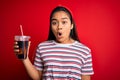 Young asian girl drinking cola fizzy refreshment using straw over isolated red background scared in shock with a surprise face, Royalty Free Stock Photo