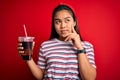 Young asian girl drinking cola fizzy refreshment using straw over isolated red background serious face thinking about question, Royalty Free Stock Photo